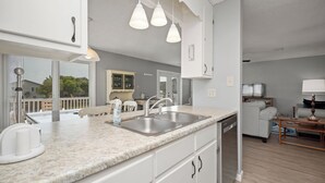 Uma cozinha moderna com bancada de granito, pia de aço inoxidável e armários brancos. É possível ver uma área de jantar com mesa e cadeiras através de uma porta aberta.