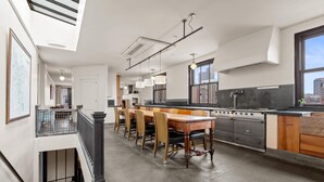 Een moderne keuken met een eethoek, met een lange houten tafel en stoelen, hangende lampen en een groot raam dat uitzicht biedt op de stad.