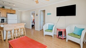 A modern kitchen with wooden cabinets, a white refrigerator, and a bar area with stools. There is a ceiling fan, a flat-screen TV mounted on the wall, and wicker chairs with cushions. - The Hideaway: Dog-friendly Unit in Multiplex Close to Public Beach Access (Oak Island)