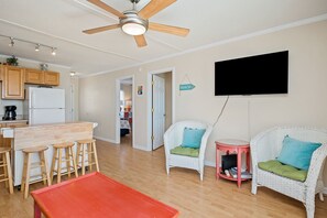 A modern kitchen with wooden cabinets, a white refrigerator, and a bar area with stools. There is a ceiling fan, a flat-screen TV mounted on the wall, and wicker chairs with cushions. - The Hideaway: Dog-friendly Unit in Multiplex Close to Public Beach Access (Oak Island)