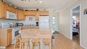 A modern kitchen with wooden cabinets, a central island with stools, a microwave, oven, and a refrigerator. - The Hideaway: Dog-friendly Unit in Multiplex Close to Public Beach Access (Oak Island)