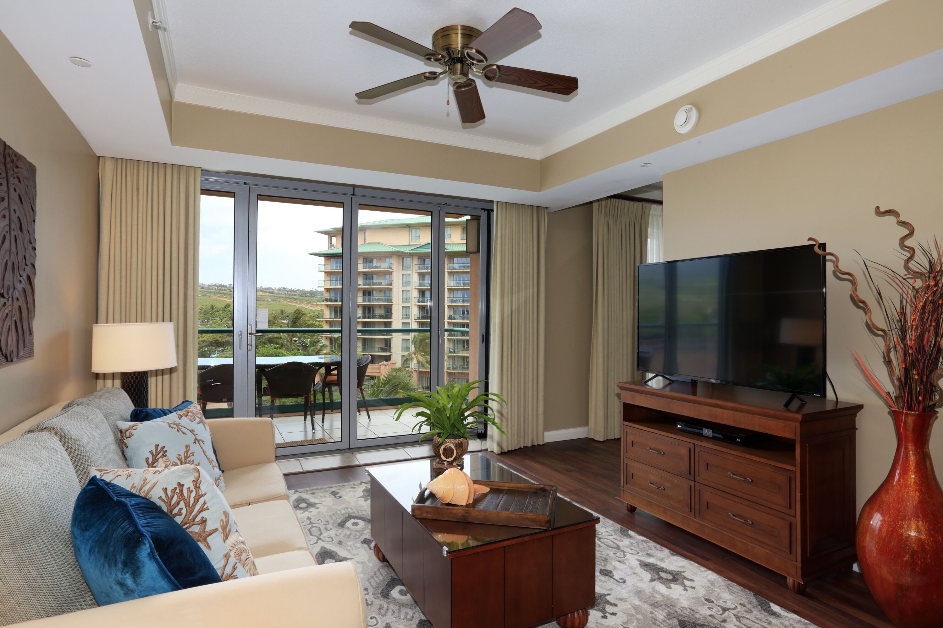 A modern living room with a sofa, coffee table, TV, and a view of a balcony with outdoor seating.