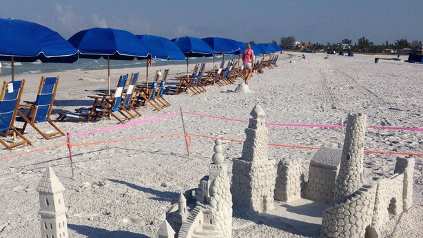 A sandy beach with a large sandcastle structure, including towers and arches, and several blue beach chairs and umbrellas in the background. - Decompress and Revitalize! Lose yourself in Paradise! (Fort Myers Beach)