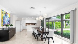 Una cocina moderna con armarios blancos, un comedor con mesa de madera oscura y sillas blancas, y un ventanal con vistas a la vegetación.