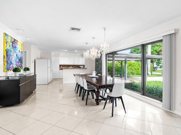 A modern kitchen with white cabinets, a dining area with a dark wooden table and white chairs, and a large window offering a view of greenery.