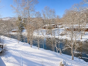 Paesaggio innevato con un fiume, alberi spogli e casette di legno.