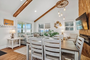 A spacious living area with a wooden ceiling, a dining table with chairs, a sofa, and a television mounted on the wall. - Into the Mystic: HGTV Renovated, Family-friendly, Oceanfront Home (Oak Island)