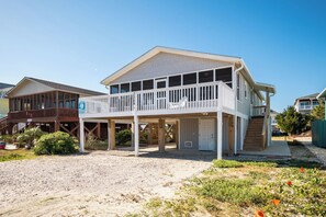 Une maison de plage à deux étages, dotée d’un balcon, d’une véranda et d’une allée menant à la maison recouverte de sable.