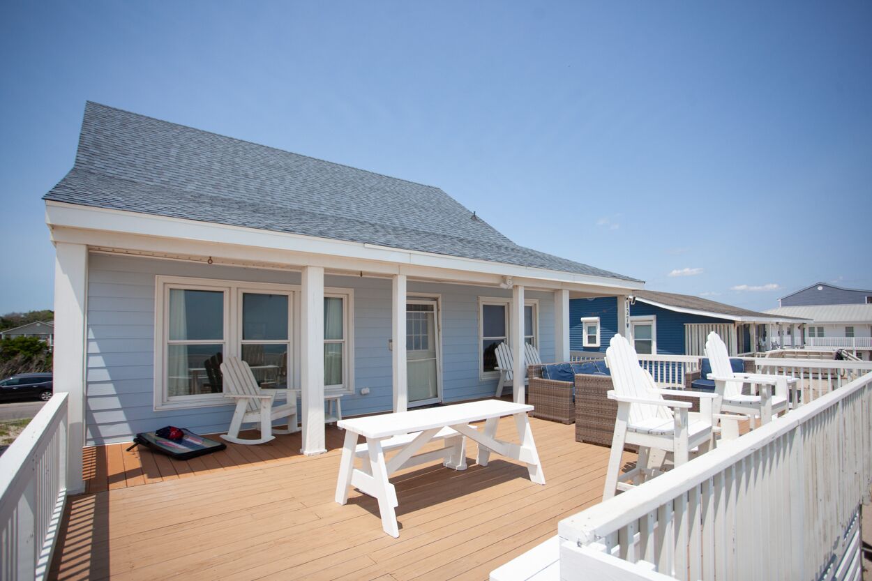 A coastal house with a wooden deck, white railings, and a covered porch with white chairs.