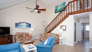 Un salon avec un canapé bleu, un divan à motifs floraux, un téléviseur et un escalier menant à un étage supérieur.