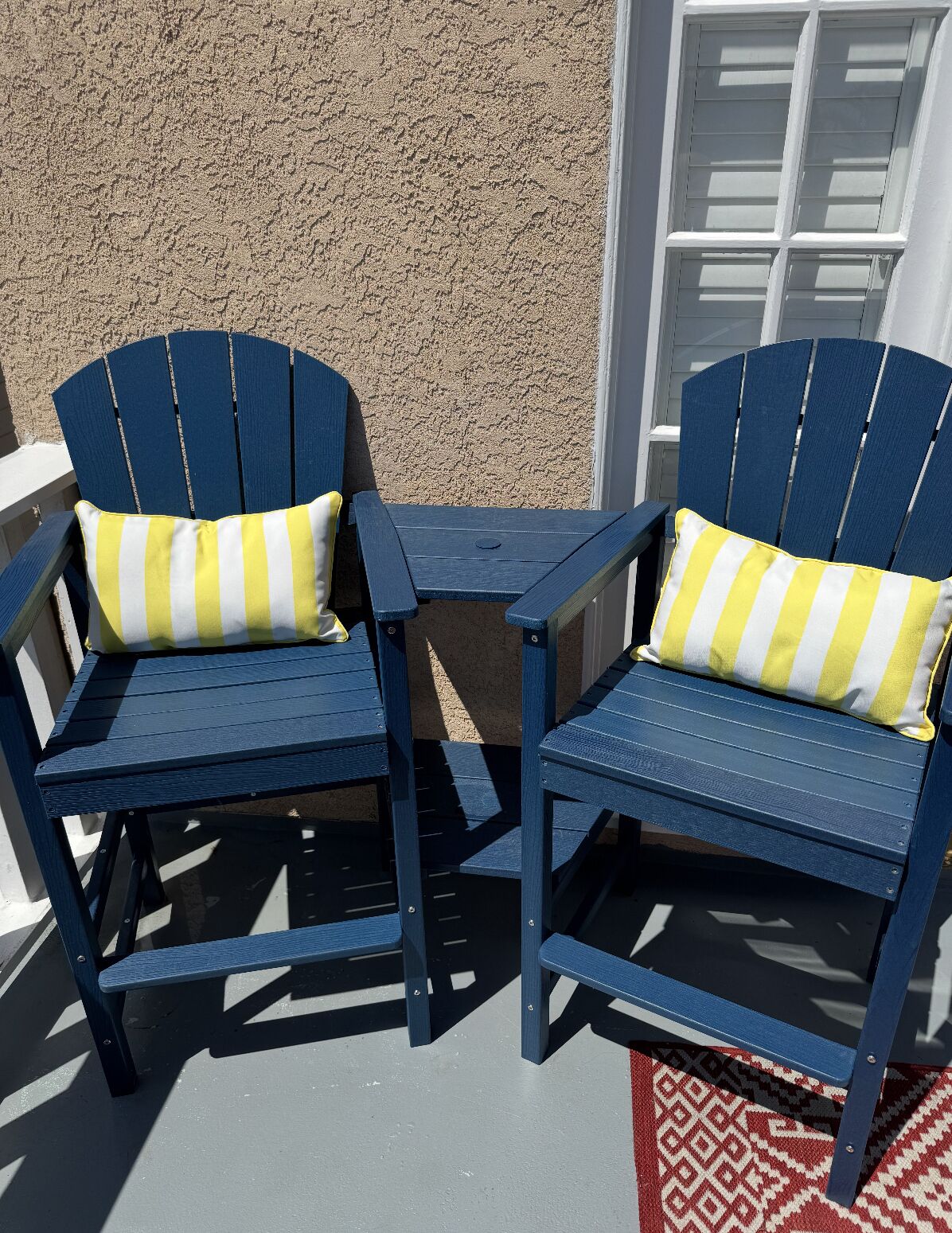 Two blue Adirondack chairs with yellow and white striped cushions, positioned on a patio with a textured wall and a window in the background.
