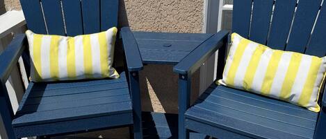 Two blue Adirondack chairs with yellow and white striped cushions, positioned on a patio with a textured wall and a window in the background.