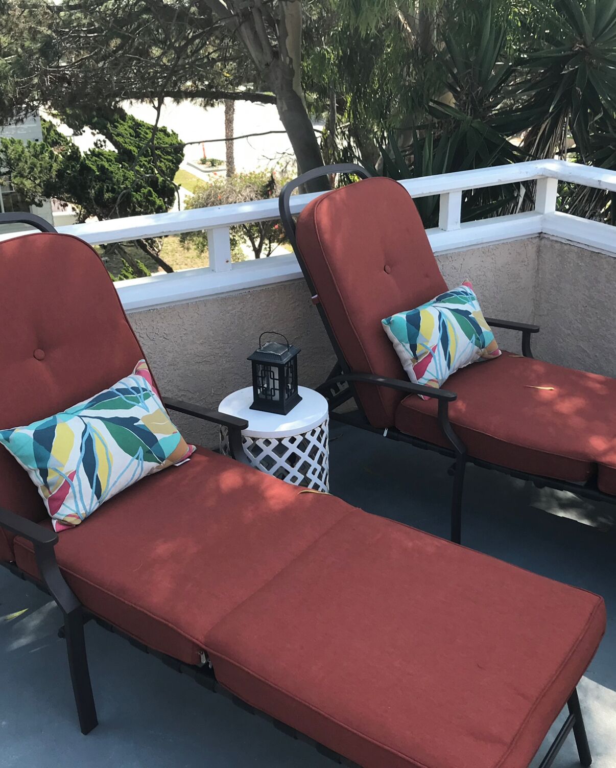 Two red lounge chairs with colorful cushions on a balcony with a white railing, a small round table with a lantern, and a view of trees and a building in the background.