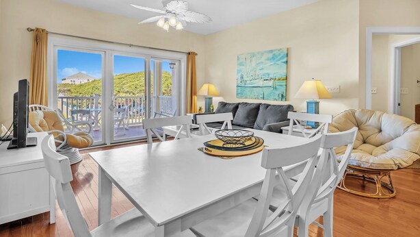 A spacious living area with a dining table, white chairs, a sofa, and a large window offering a view of a hillside. - The Dunes: Ocean View Condo with Community Pool, Hot Tub, Beach Access (Oak Island)