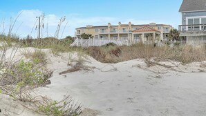 A beachfront property with a multi-story building, a covered walkway, and a sandy beach area. - The Dunes: Ocean View Condo with Community Pool, Hot Tub, Beach Access (Oak Island)