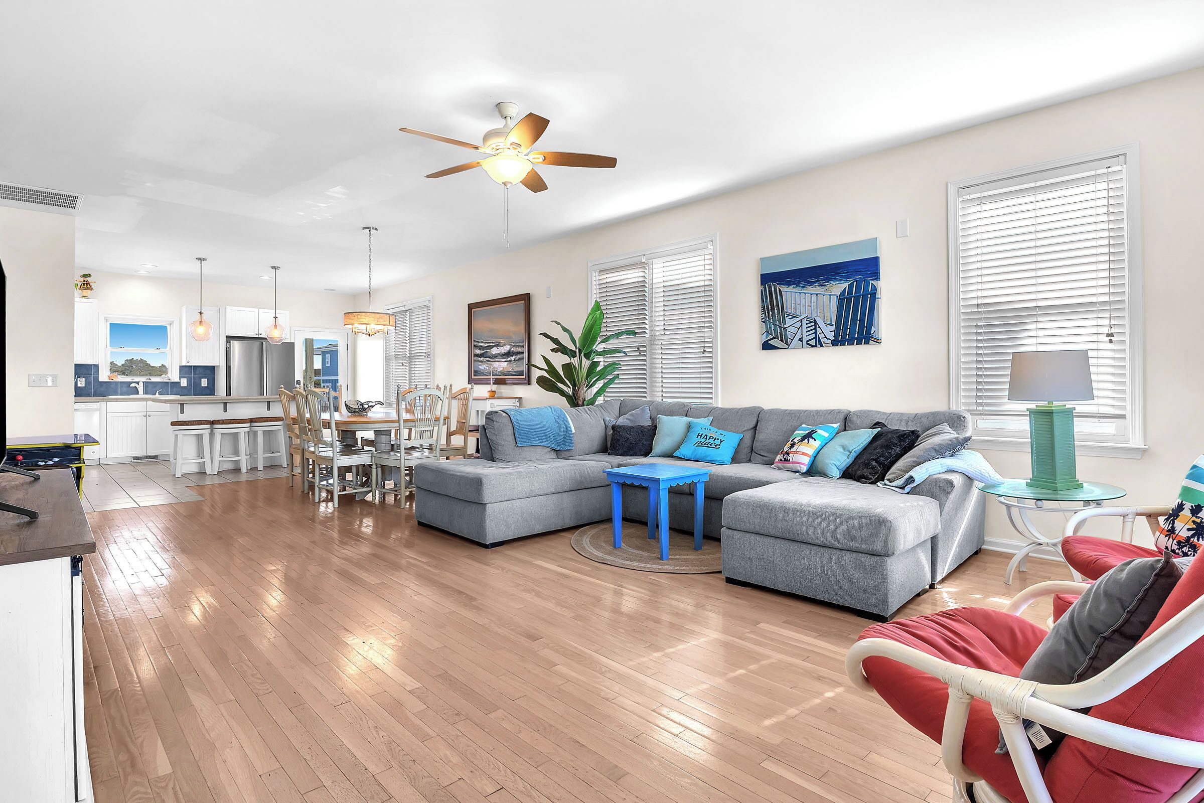 A modern living room with a grey sectional sofa, a red armchair, and a blue coffee table. There is a ceiling fan, a dining area with a table and chairs, and a kitchen in the background.
