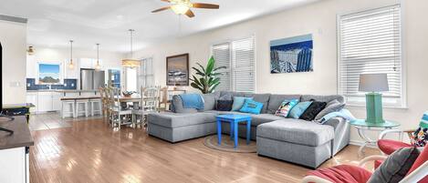 A modern living room with a grey sectional sofa, a red armchair, and a blue coffee table. There is a ceiling fan, a dining area with a table and chairs, and a kitchen in the background.