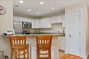 A kitchen with white cabinets, a stainless steel refrigerator, a microwave, and a wooden island with two chairs. - Dreamer: Oceanview Condo w/Pool, Hot Tub, and Elevator Access (Oak Island)