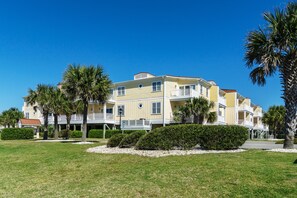 A multi-story hotel with a flat roof, balconies, and a well-maintained lawn with palm trees.