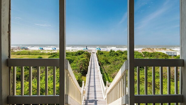 Una escalera de madera que lleva a una playa con vistas claras al océano y dunas lejanas.