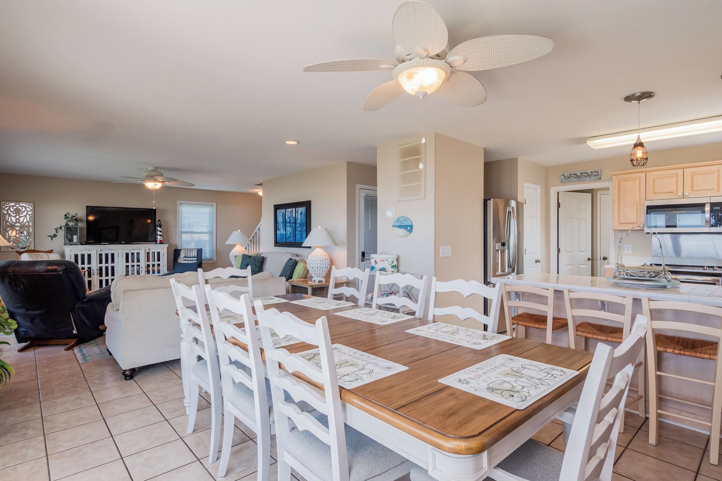 A spacious dining area with a wooden table and white chairs, a ceiling fan, and a kitchen in the background.