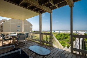 A wooden deck with outdoor furniture, a view of the ocean, and a building in the background. - Sweet Retreat: Oceanfront Home with Private Walkway to the Beach (Oak Island)