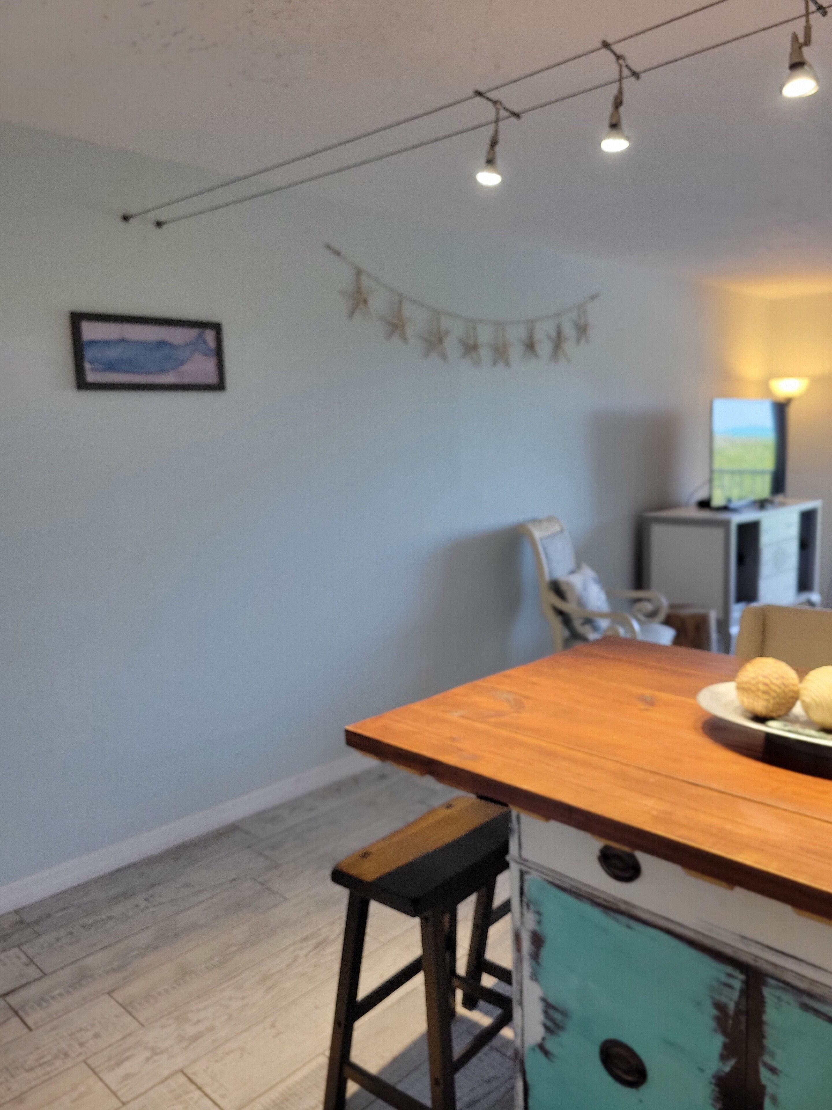 A dining area with a wooden table, two barstools, a wall-mounted picture, string lights, and a small cabinet.