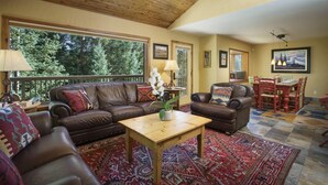 A spacious living room with a wooden ceiling, large windows, a brown leather sofa, a wooden coffee table, and a dining area with a red table and chairs. - Blue Bend Beauty | Comfort & Privacy with Postcard Views & Private Hot Tub (Breckenridge)