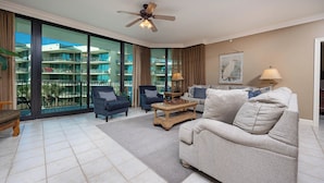 A spacious living room with a large window, a ceiling fan, and a view of the building's exterior balconies. - BRING the BOAT!! 3 BR 3 BA…Phoenix On The Bay!! (Orange Beach)