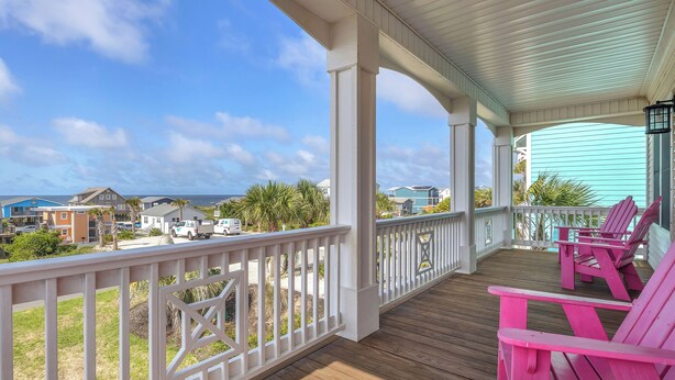 Een overdekt terras met een houten dek, witte relingen en roze Adirondack-stoelen.