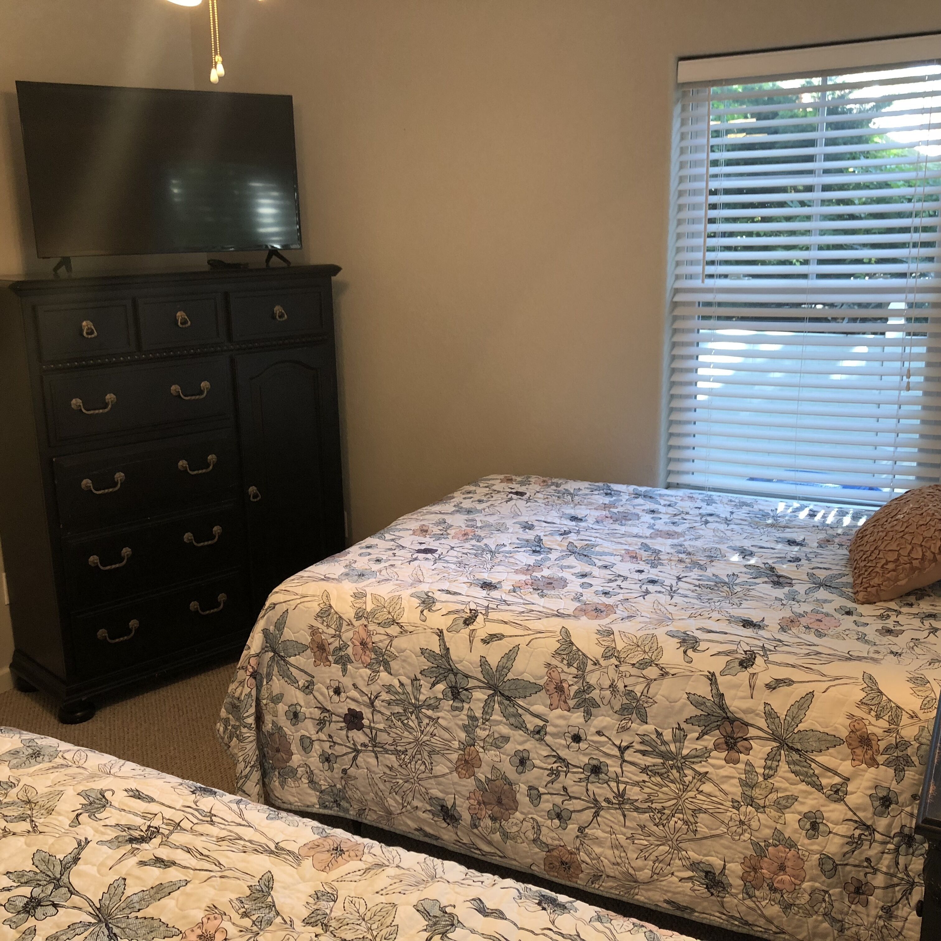 Une chambre à coucher avec un lit, une commode, un téléviseur posé sur la commode et une fenêtre avec des stores.
