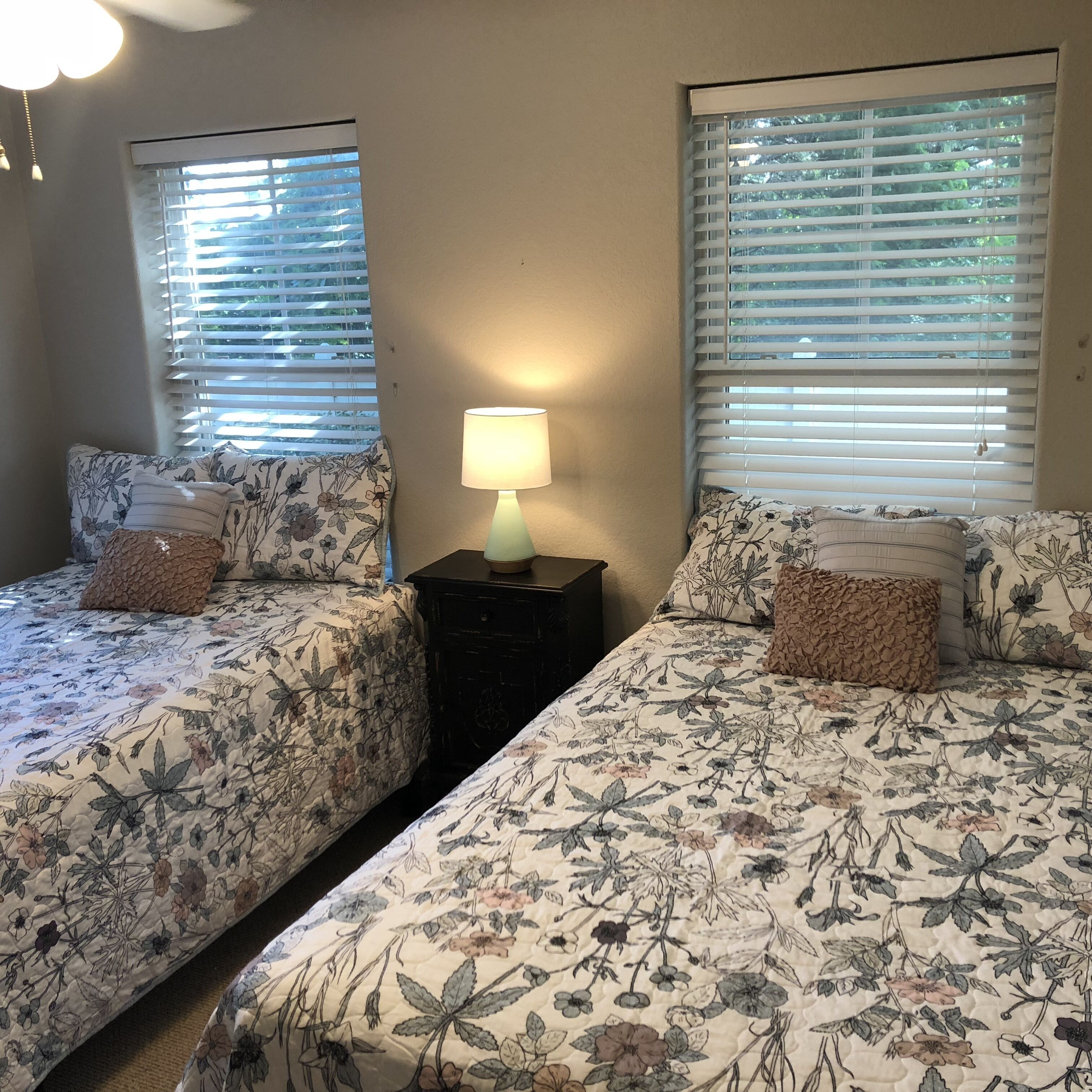 A bedroom with two beds, each with floral bedding, a bedside table with a lamp, and windows with blinds.