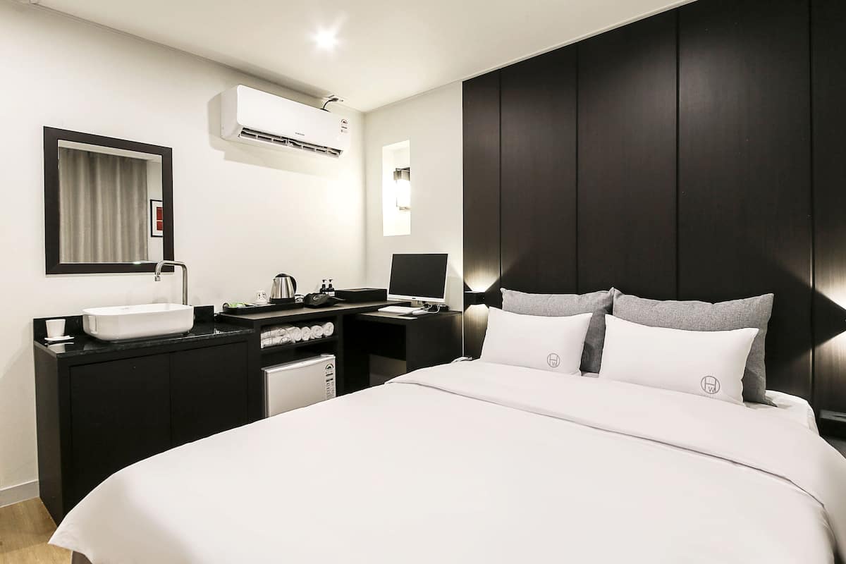 A modern hotel room with a large bed, a bedside table, a built-in desk, a vanity with a mirror, and an air conditioning unit.