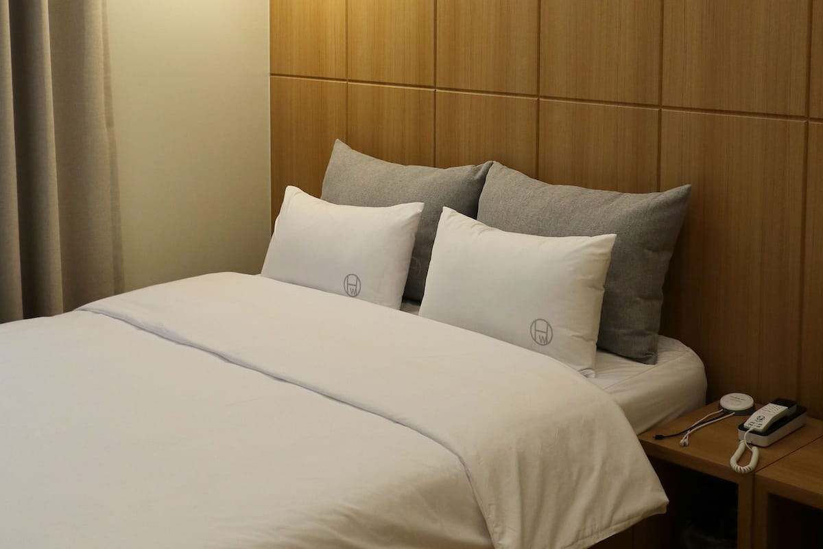 A neatly made bed with white and grey pillows, a wooden headboard, and a bedside table with a phone and charger.