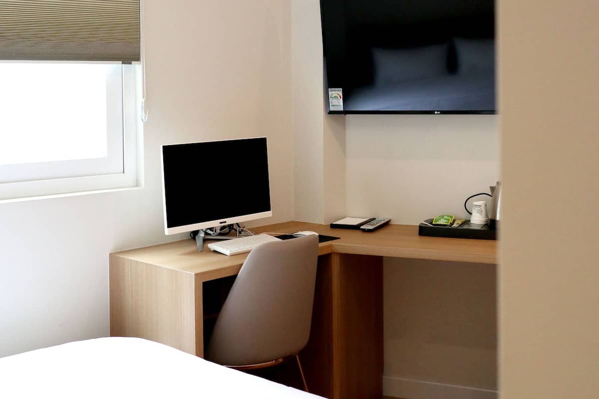 A hotel room with a bed, a desk with a computer, a chair, a TV mounted on the wall, and a window with blinds.
