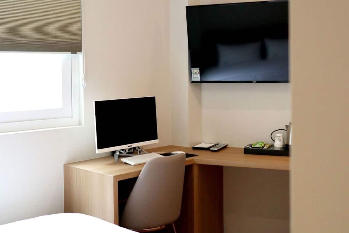 A hotel room with a bed, a desk with a computer, a chair, a TV mounted on the wall, and a window with blinds.