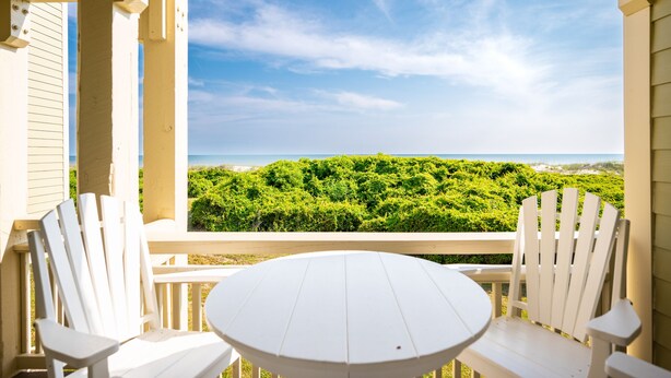 A covered outdoor seating area with a round table and two white Adirondack chairs, overlooking a lush green landscape and the ocean. - Garris Hideaway 2: Cozy 1st Level Oceanfront Condo - Perfect for Couples Getaway (Caswell Beach)