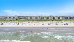 A beachfront hotel with multiple buildings, a sandy beach with umbrellas, and waves in the foreground. - Garris Hideaway 2: Cozy 1st Level Oceanfront Condo - Perfect for Couples Getaway (Caswell Beach)