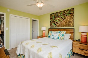 A bedroom with a bed, two bedside tables, a ceiling fan, a door, and a wall with a leaf-patterned painting.