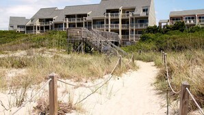 A beachfront hotel with multiple balconies, a sandy path leading to the entrance, and a wooden staircase connecting the buildings. - Garris Hideaway 2: Cozy 1st Level Oceanfront Condo - Perfect for Couples Getaway (Caswell Beach)