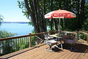 Eine Holzterrasse mit Tisch, Stühlen und einem Sonnenschirm, mit Blick aufs Wasser.