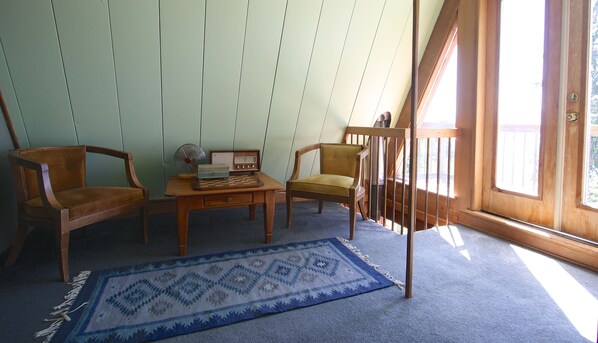 Ein Zimmer mit einem Holzbüro, zwei Stühlen, einem kleinen Tisch mit Schachbrett, einem Radio und einem blau gemusterten Teppich.