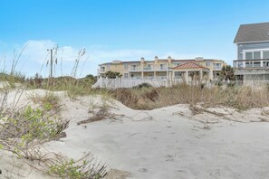 A beachfront property with a two-story building, a covered patio, and a sandy beach area. - Star Fish: Ocean View Condo with Pool and Hot Tub (Oak Island)