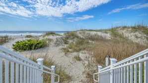 Ein Strand mit einer weißen Treppe, die zum Sand hinabführt, ein hölzerner Steg im Hintergrund und ein klarer, blauer Himmel.