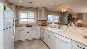 A kitchen with white cabinets, a stone backsplash, a window with blinds, a ceiling fan, and a countertop with a toaster and a dish rack. - Fish Tales: Charming Oceanfront Condo with Wrap-around Deck, Central Location Id (Oak Island)