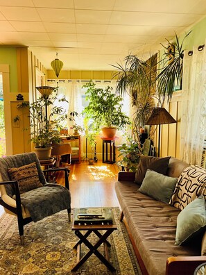 Una sala de estar acogedora con sofá, sillón y una mesita de centro. Hay varias plantas en macetas y una lámpara.