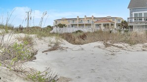 Una proprietà fronte spiaggia con un edificio a più piani, una galleria coperta e terreno sabbioso con vegetazione rada.
