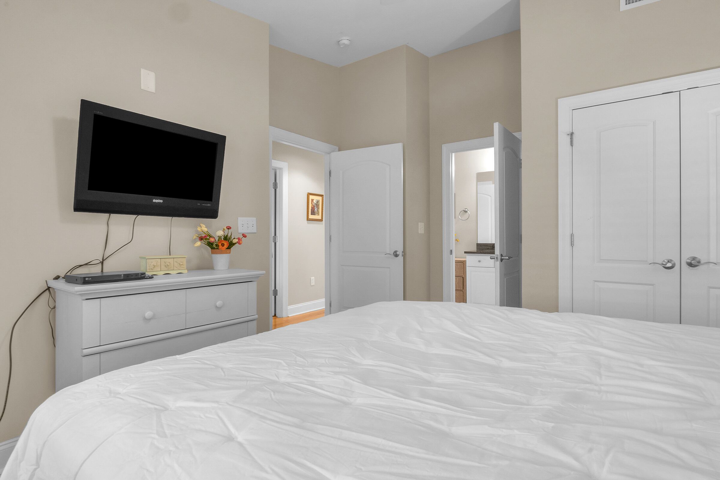 Une chambre dotée d’un grand lit, d’un téléviseur à écran plat fixé au mur, d’une commode et d’une porte donnant sur une autre pièce.