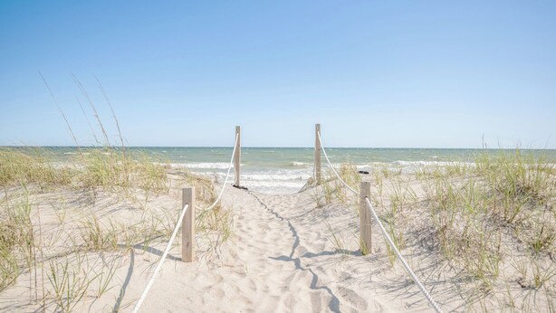 Un sentier de sable qui longe la plage et mène à l’océan, bordé de poteaux et de cordes en bois.