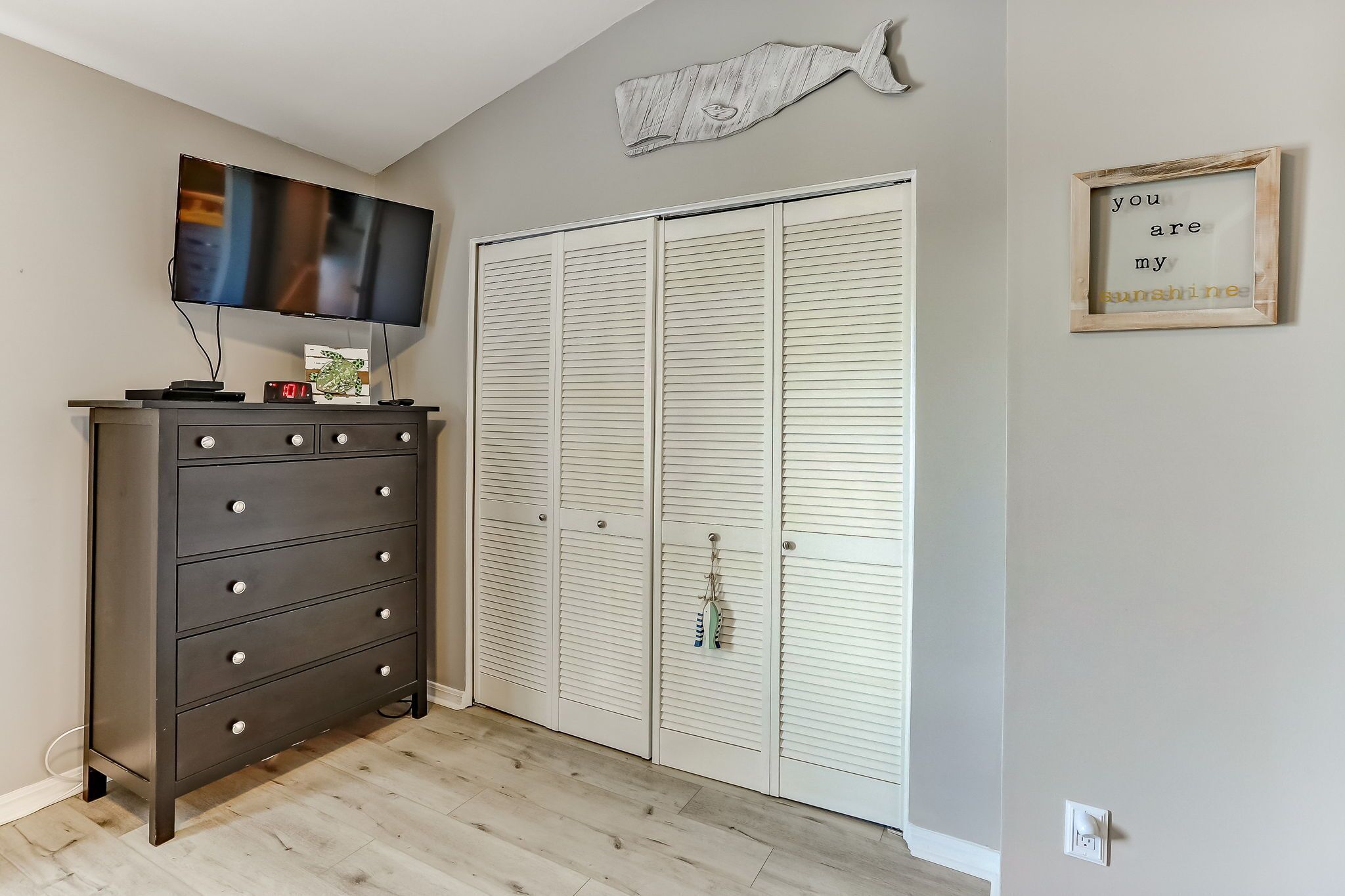 A room with a dark wooden dresser, a television, a white closet with louvered doors, and a wall-mounted fish decoration.